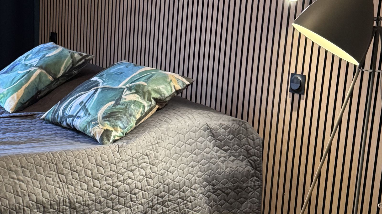Hotellihuone illalla, sänky ja lukuvalo puusäleseinää vasten